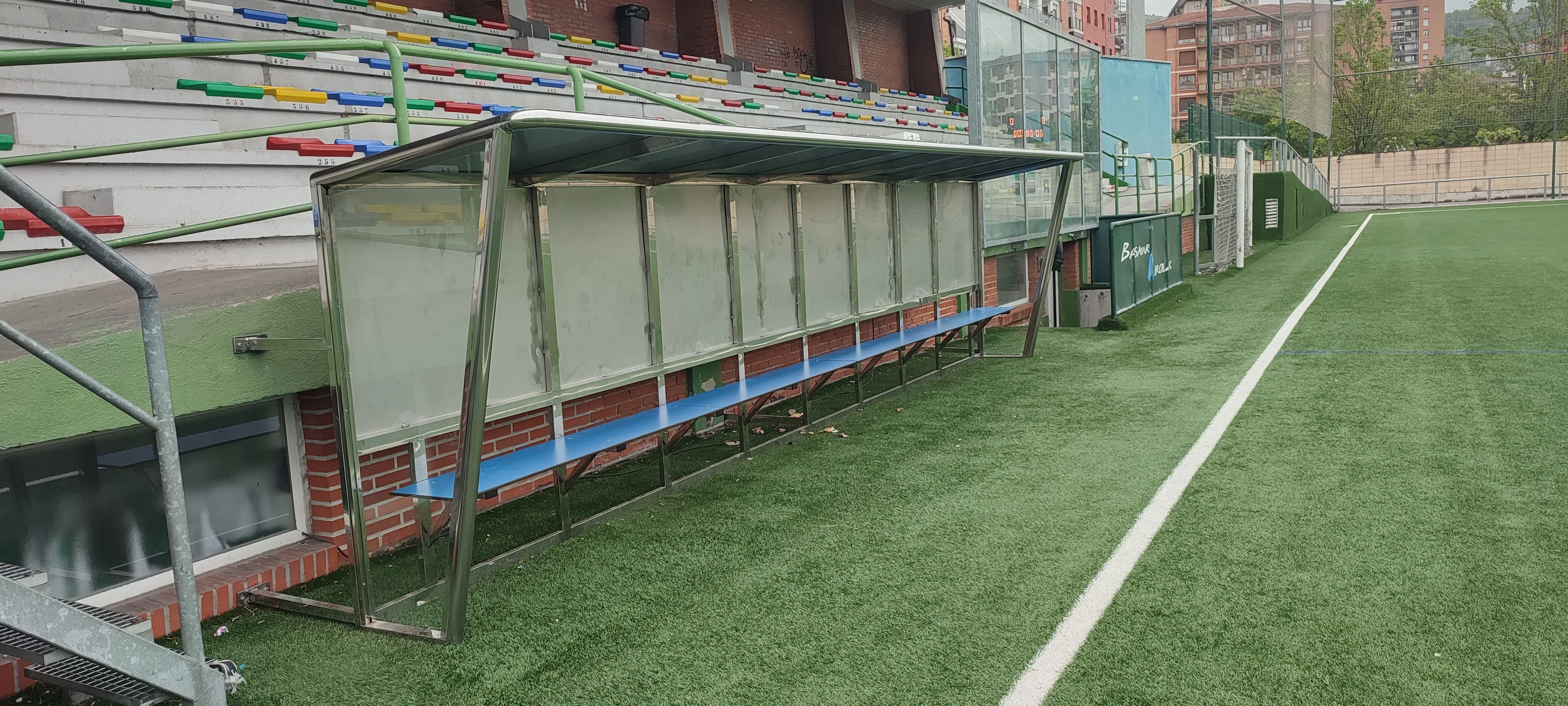 Banquillo largo con estructura metálica y cubierta junto a un campo de césped artificial