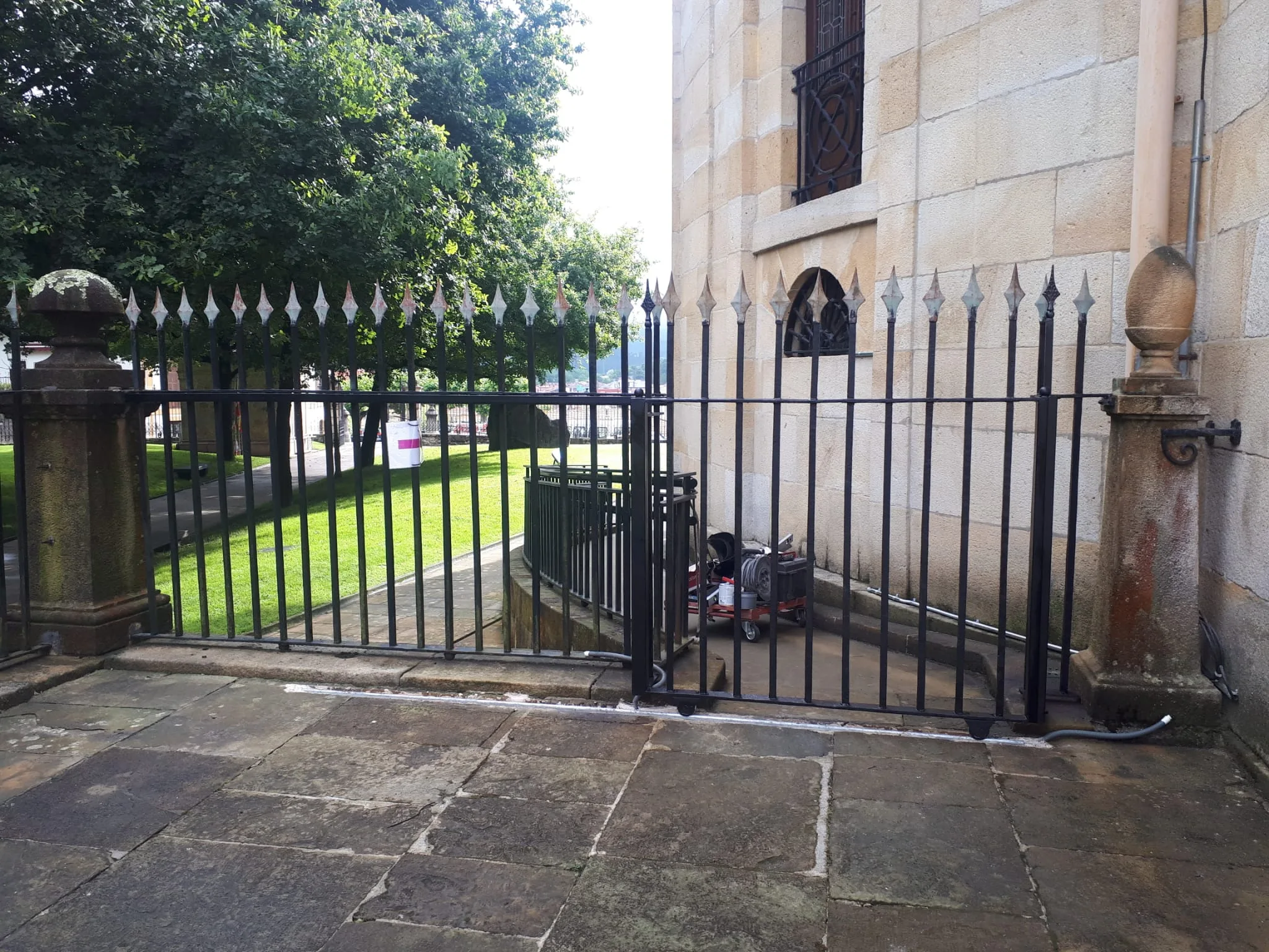 Puerta de herrería negra con barrotes verticales junto a un edificio de piedra