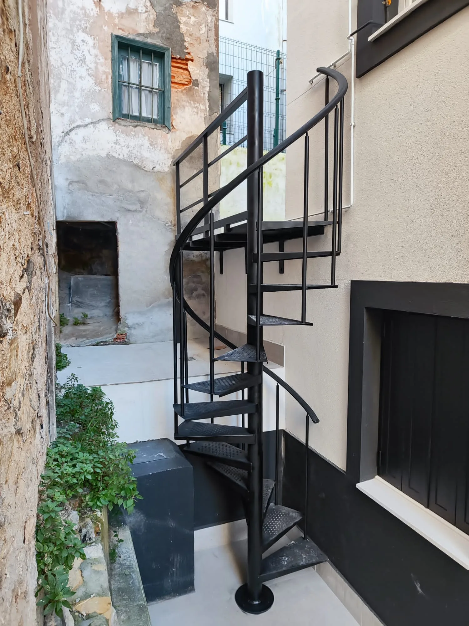 Escalera de caracol negra instalada entre una pared de piedra y una fachada blanca