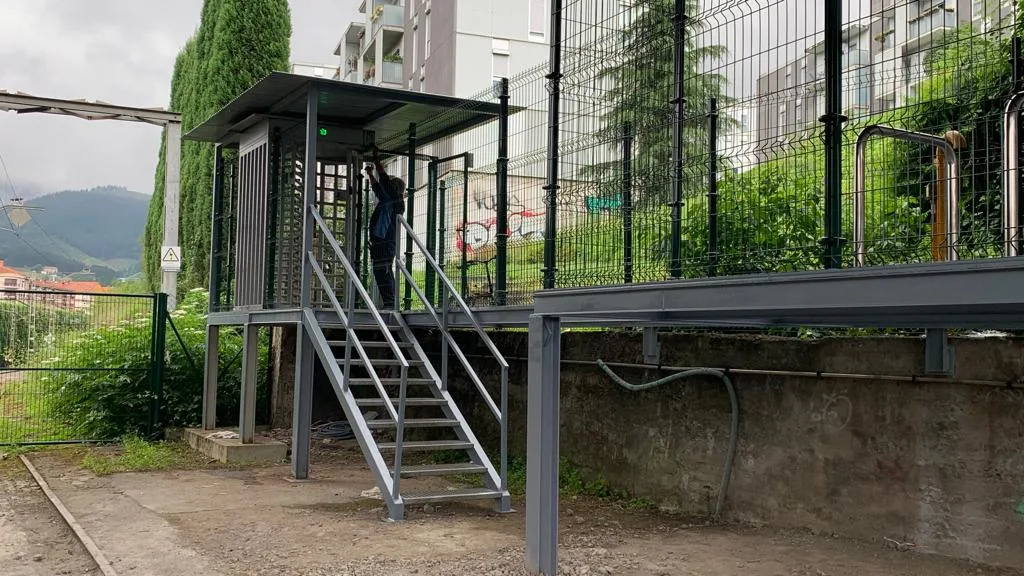 Plataforma metálica gris con escalera y caseta cubierta junto a un vallado verde
